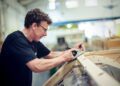 A man uses a hand plane to smooth a wooden board in a workshop, crafting details reminiscent of the Morgan Plus Four 75th Anniversary’s timeless design.