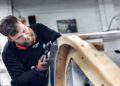 A man uses a power tool to work on the metal frame of a wooden structure in a workshop, wearing ear protection around his neck and gloves on his hands—capturing the craftsmanship behind the Morgan Plus Four 75th Anniversary.