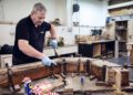 A man in a workshop uses clamps and tools to work on a curved wooden structure for a Morgan Plus Four, wearing blue gloves and a black shirt. Various tools and materials are scattered on the table.