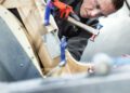 A person wearing safety glasses and gloves uses a hammer to work on a wooden and metal structure, with clamps holding materials in place—a nod to the craftsmanship behind the Morgan Plus Four 75th Anniversary.