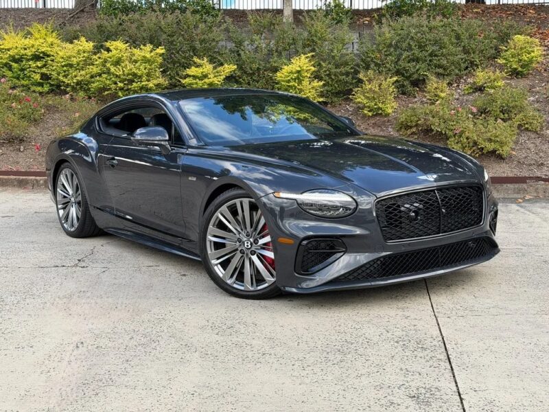 A dark gray luxury coupe with large alloy wheels, this exotic car is parked on a concrete driveway in front of lush shrubbery.
