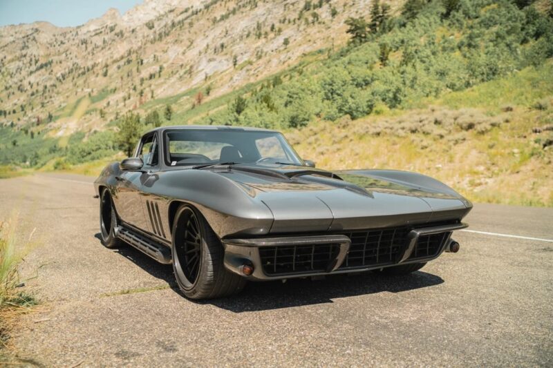 A classic black Chevrolet Corvette, one of the quintessential Classic Muscle Cars, is parked on a mountain road with trees and rocky hills in the background.
