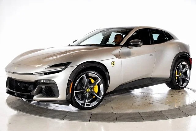 A silver Ferrari SUV, an exotic car with yellow brake calipers, is parked indoors on a circular platform, viewed from the front left side.