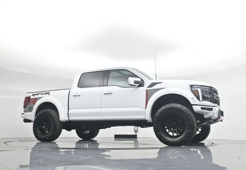 A white Ford F-150 Raptor pickup truck, known for its comfort and style, is parked in a studio with a plain white background, viewed from the side.