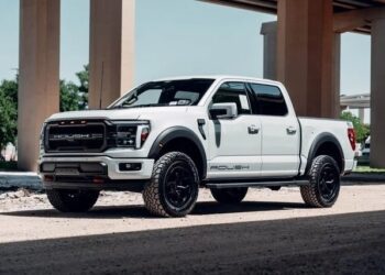 A white Roush-modified Ford F-150 pickup truck, blending luxury, comfort, and style, is parked on a gravel surface under an overpass, with trees and pillars in the background.
