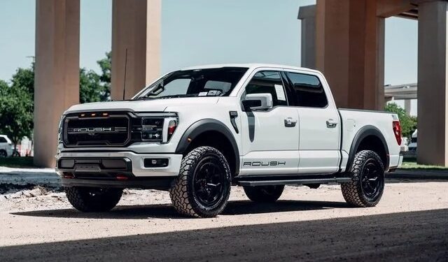 A white Roush-modified Ford F-150 pickup truck, blending luxury, comfort, and style, is parked on a gravel surface under an overpass, with trees and pillars in the background.