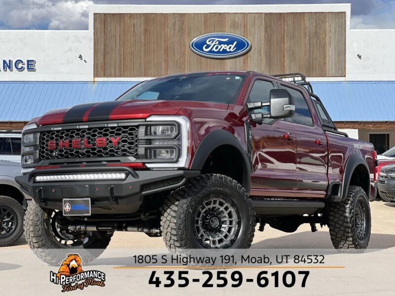 A lifted red Ford Shelby pickup truck with black stripes, known for its luxury trucks comfort and capability, is parked in front of a Ford dealership in Moab, Utah. Contact details and address are displayed at the bottom.
