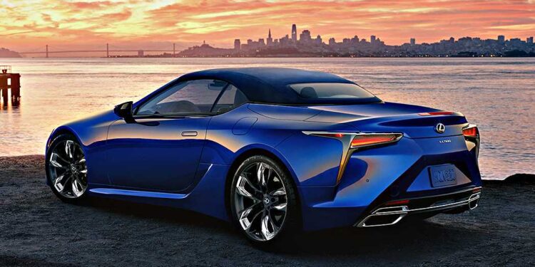 A blue Lexus sports car—featured in our Review: 2025 Lexus LC 500 Convertible—parked by the water at sunset, with a city skyline and bridge visible in the background.