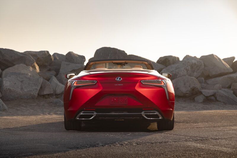 Review: 2025 Lexus LC 500 Convertible—a striking red model is parked on a paved area near large rocks, shown from the rear, basking in soft sunlight.