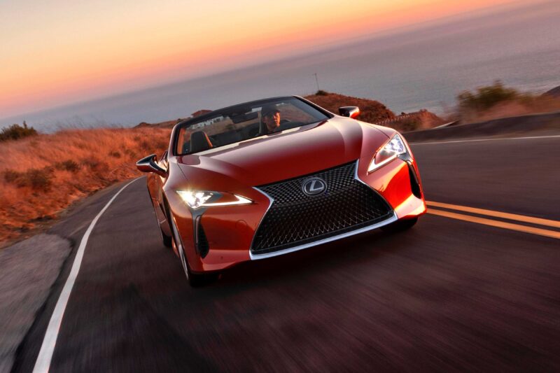 A red Lexus convertible cruises along a curving coastal road at sunset, ocean and dry grass in the background. Review: 2025 Lexus LC 500 Convertible captures this thrilling drive with breathtaking scenery.