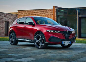 A red 2026 Alfa Romeo Tonale hybrid SUV is parked on a stone driveway in front of a modern brick and glass building.