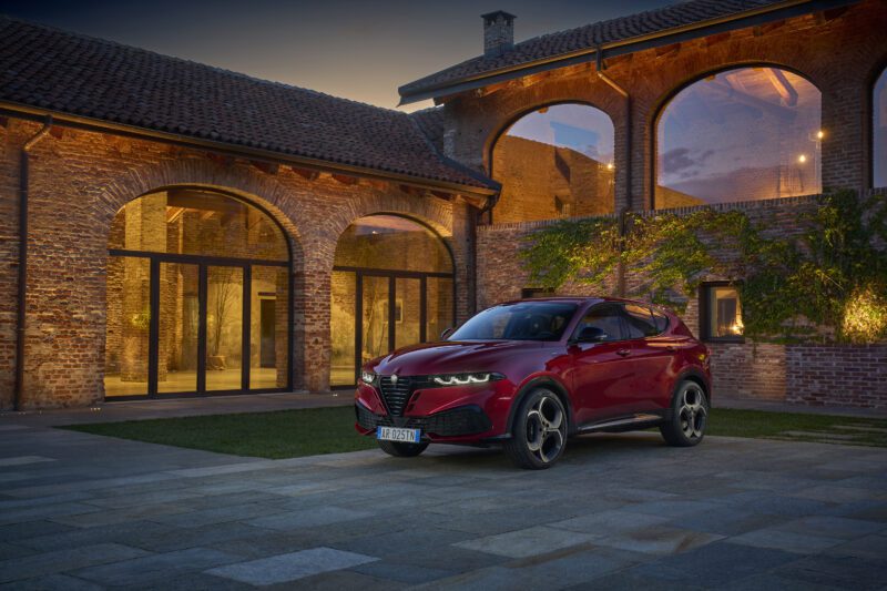 A red Alfa Romeo Tonale SUV, featuring hybrid power, is parked in front of a brick building with large arched windows in the evening.