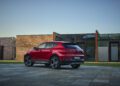 A red Alfa Romeo Tonale SUV with hybrid power is parked on a stone driveway in front of modern brick and glass buildings, under a clear sky.