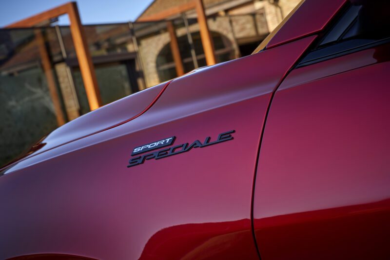 Close-up of a red Alfa Romeo Tonale’s side panel showing the “Sport Speciale” badge, with a building and glass panels blurred in the background—an updated look for 2026.