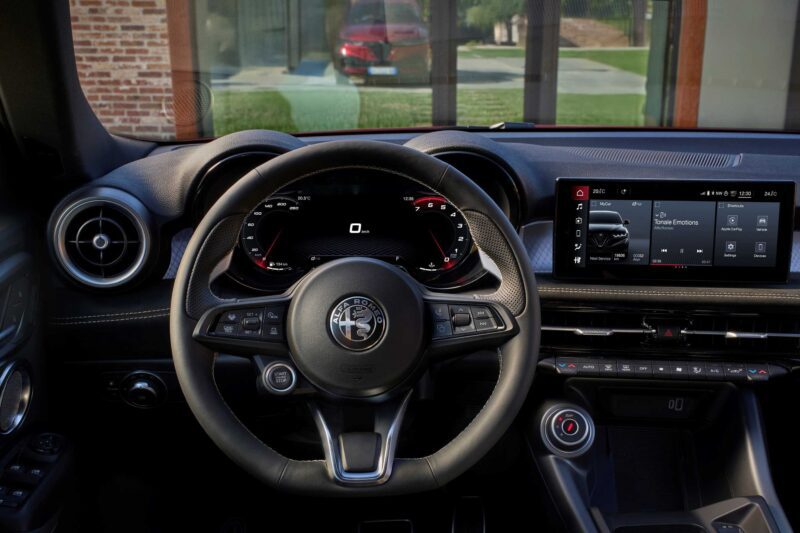 A view of a modern car interior showing the steering wheel with the Alfa Romeo logo, a digital instrument cluster, and a center touchscreen display—highlighting the 2026 Alfa Romeo Tonale with advanced hybrid power.