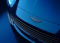 Close-up view of the front grille and emblem of a blue Aston Martin DB12 S Super Tourer, photographed under blue lighting.