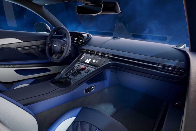 Interior view of an Aston Martin DB12 S Super Tourer featuring a digital dashboard, touchscreen controls, a multifunction steering wheel, and blue ambient lighting.