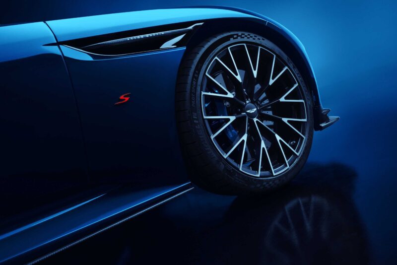 Close-up of a blue Aston Martin DB12 S sports car’s front wheel and fender, featuring a multi-spoke alloy rim, red “S” badge, and sharp bodywork—capturing the bold design of modern super tourers against a blue background.