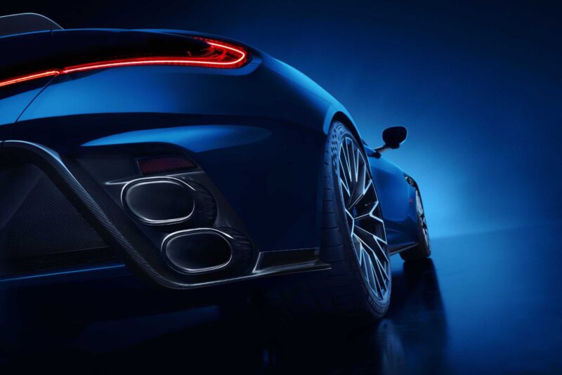 Close-up of the rear section of a blue Aston Martin DB12 S Super Tourer, highlighting the taillight, dual exhaust pipes, and large wheel against a striking blue gradient background.