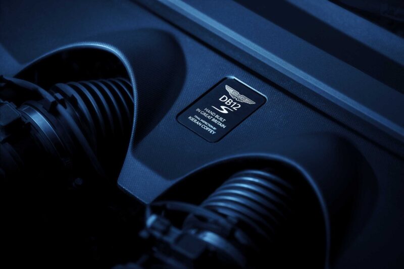 Close-up view of an Aston Martin DB12 S engine bay with a badge reading "Hand Built in England" and engine components visible, showcasing the craftsmanship behind the Super Tourers lineup.