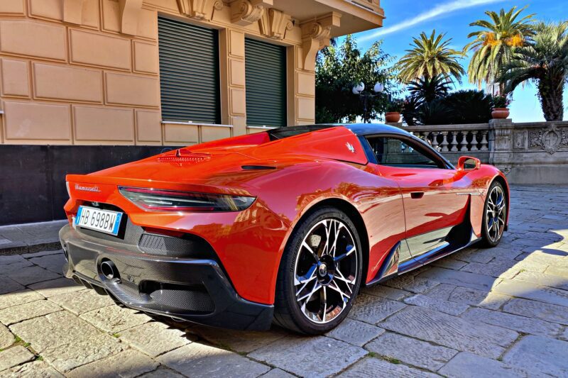 A red 2026 Maserati MCPura Cielo First Drive sports car is parked on a stone street beside a beige building, with palm trees and a blue sky in the background.