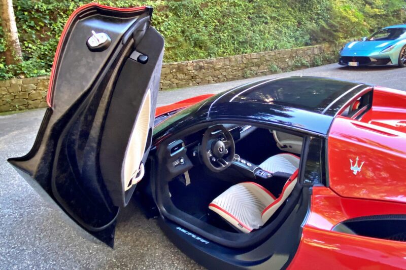 A 2026 Maserati MCPura Cielo First Drive sports car in red sits with its driver-side scissor door open, revealing a striking black and white interior; another car is parked in the background.