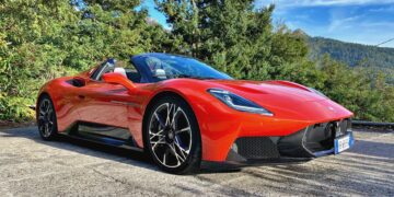 A 2026 Maserati MCPura Cielo First Drive red convertible sports car is parked on a concrete surface near trees and mountains under a blue sky.
