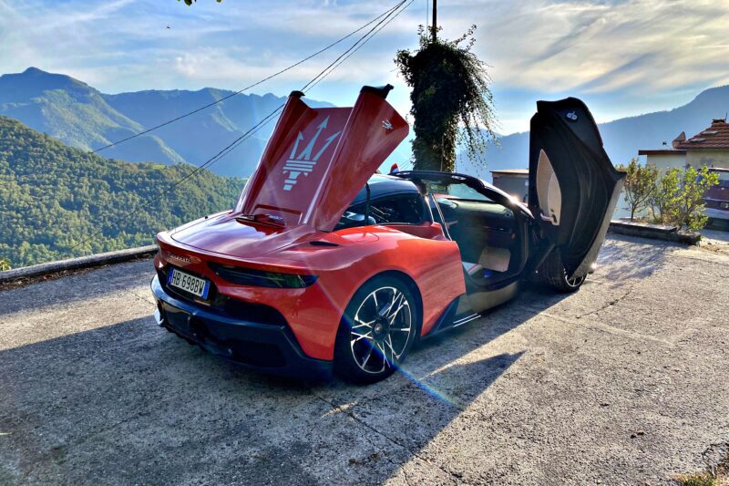 A red 2026 Maserati MCPura Cielo First Drive sports car with its butterfly doors open is parked on a mountain road, framed by scenic hills and a house in the background.