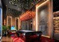 A modern game room at Aureum Estate with a pool table, geometric ceiling panels, a red rug, a large chandelier, indoor plants, and wall-mounted racks for cues and wine bottles, inspired by the luxury of Dubai Hills.
