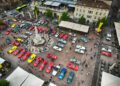 Aerial view of a city square with rows of colorful vintage cars on display, surrounded by crowds of people and outdoor cafes.