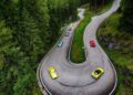 A group of colorful sports cars, including those from the Ferrari Cavalcade, drive along sharp curves on a winding mountain road in South Tyrol, Italy, surrounded by dense green forest.