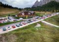 A large group of classic sports cars, including stunning Ferraris from the Cavalcade Classiche, is parked around a lodge in South Tyrol’s mountainous, forested area under a cloudy sky.