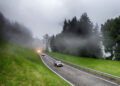 Several cars, including participants of the Ferrari Cavalcade, drive along a winding road surrounded by green grass and trees in South Tyrol on a foggy day.