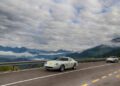 A classic silver sports car with race decals drives on a mountain road during the Ferrari Cavalcade in South Tyrol, Italy, followed by another car, with clouds and mountains visible in the background.