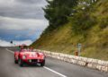 A red vintage race car with the number 437 drives along a winding mountain road in South Tyrol, Italy, lined with grass, trees, and a stone barrier under a cloudy sky—echoing the spirit of the Ferrari Cavalcade.