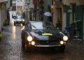 Several classic cars, including those from the Ferrari Cavalcade, drive on a narrow, wet cobblestone street lined with old buildings in South Tyrol, Italy during rainy weather.