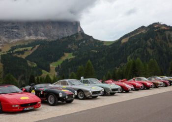 A row of classic and vintage sports cars, part of the Ferrari Cavallcade, parked by a mountain road in South Tyrol, Italy, with forested hills and cloudy skies in the background.