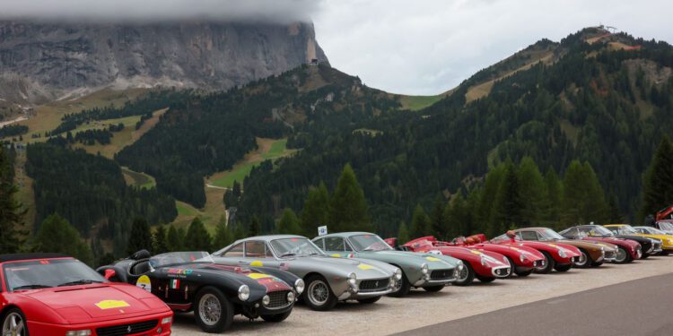 A row of classic and vintage sports cars, part of the Ferrari Cavallcade, parked by a mountain road in South Tyrol, Italy, with forested hills and cloudy skies in the background.