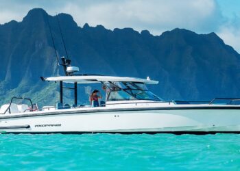 A white motorboat with two people on board floats on turquoise water, offering a true maritime experience, with a lush green mountain range in the background under a partly cloudy sky.