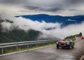Two vintage sports cars drive along a winding mountain road in South Tyrol, with low clouds and forested hills in the background—capturing the spirit of Italy and the elegance of Ferrari Cavalcade Classiche.