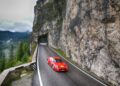 A red sports car drives on a mountain road in South Tyrol, Italy, passing through a rock tunnel with pine trees and distant mountains under a cloudy sky, capturing the spirit of the Ferrari Cavalcade.