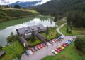 Aerial view of a lakeside building in South Tyrol, surrounded by trees, with rows of colorful Ferrari sports cars from the Cavalcade Classiche event parked outside and people gathered near the vehicles.