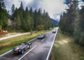 Three sports cars drive along a two-lane road through a forested, mountainous landscape in South Tyrol, Italy, under a partly cloudy sky.