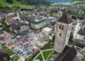 Aerial view of a small South Tyrol town in Italy with clusters of colorful cars, including those from the Ferrari Cavalcade, parked in a central square, surrounded by charming buildings, a church tower, and rolling green hills.