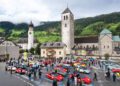 A crowd gathers around numerous sports cars in a historic European village square, as the Ferrari Cavalcade winds through South Tyrol, with charming churches, old buildings, and green hills in the background.