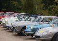 A row of classic vintage cars parked on a gravel surface in South Tyrol, Italy, with trees and greenery in the background—an elegant scene reminiscent of the Ferrari Cavalcade.