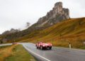 A red vintage sports car with two passengers drives on a winding mountain road in South Tyrol, Italy, with rocky cliffs and grassy hills in the background.