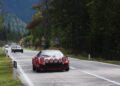 A red classic sports car, part of the Ferrari Cavalcade, drives on a two-lane road in South Tyrol, Italy, bordered by trees and road signs, with other vehicles following behind.