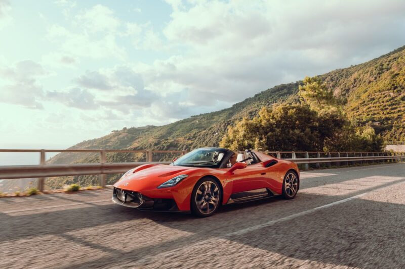 A 2026 Maserati MCPura Cielo First Drive takes a red convertible sports car along a coastal road, surrounded by hills and greenery beneath a partly cloudy sky.