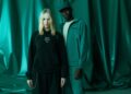 Two people stand in front of a green draped background; one wears a black hoodie and skirt, while the other sports a green jacket, pants, and cap from the PUMA Motorsport F1 Team Collection.